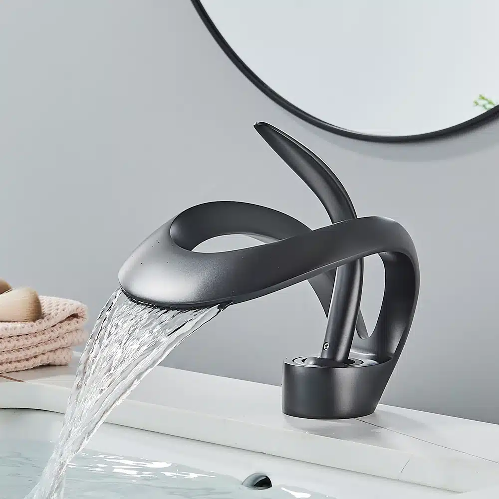 Kuldse luksusliku disainiga vannitoa segisti, millel on voolujooneline ja modernne vorm. Segisti kallab vett elegantselt valge kraanikausi kohale, taustal heleroosa volditud käterätik ja hall sein.