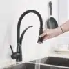 Köögivalamu kraan musta viimistlusega, pööratav väljavool | E-pood Tootemaailm Elegantne musta viimistlusega köögivalamu kraan pööratava väljavooluga, mille paindlik voolik ulatub roostevabast terasest kraanikaussi, valgel taustal. Käsi suunab kraani väljavoolikut, näidates tugevat veevoolu.