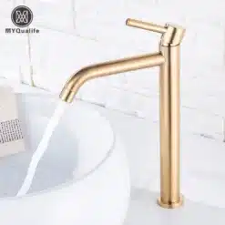 Elegantne kuldne kraan vannituppa, voolava veega, valge kraanikausi äärel. Taustaks hele tellissein, rõhutades luksuslikku viimistlust.