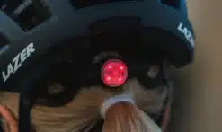 Jalgrattakiivri tagaosas asuv LED-tagatuli LAZER Light TS+ Led One on punane ja heleduses, parandades nähtavust hämaras. Tuli on kinnitatud mustale kiivrile koos nähtava logoga ja valgus paistab juuksekummiga patsi lähedal.