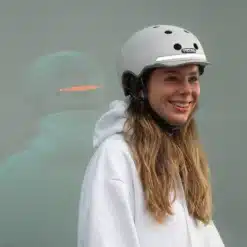 Naeratav naine kannab valget Kiiver MELON E-Series, Metropolis (M/L) kiivrit, hoides taustaks minimalistlikku heledat seina.