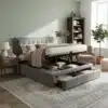 Kaheinimesevoodi panipaikade ja sahtlitega, hall 140x200 toas puidust põrandal, pehme peats ja avatud panipaik koos sahtlitega. Taustal pastelne sein, raamatukapp ja tugitool.