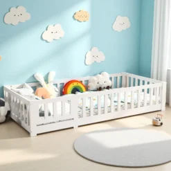 Võrega lastevoodi männipuidust 90x200 cm, valge värvusega, pastellsete pilvede kaunistustega sinisel seinal. Voodi on täidetud värviliste padjade ja pehmete leludega, asub valgusküllases toas heleda puidust põrandaga.