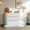 Stiilne valge kapp LED-valgustusega, millel on kaks ust ja kolm sahtlit, sobitub kaasaegsesse elutuppa heleda lambivalguse ja dekoratiivsete elementide taustal.