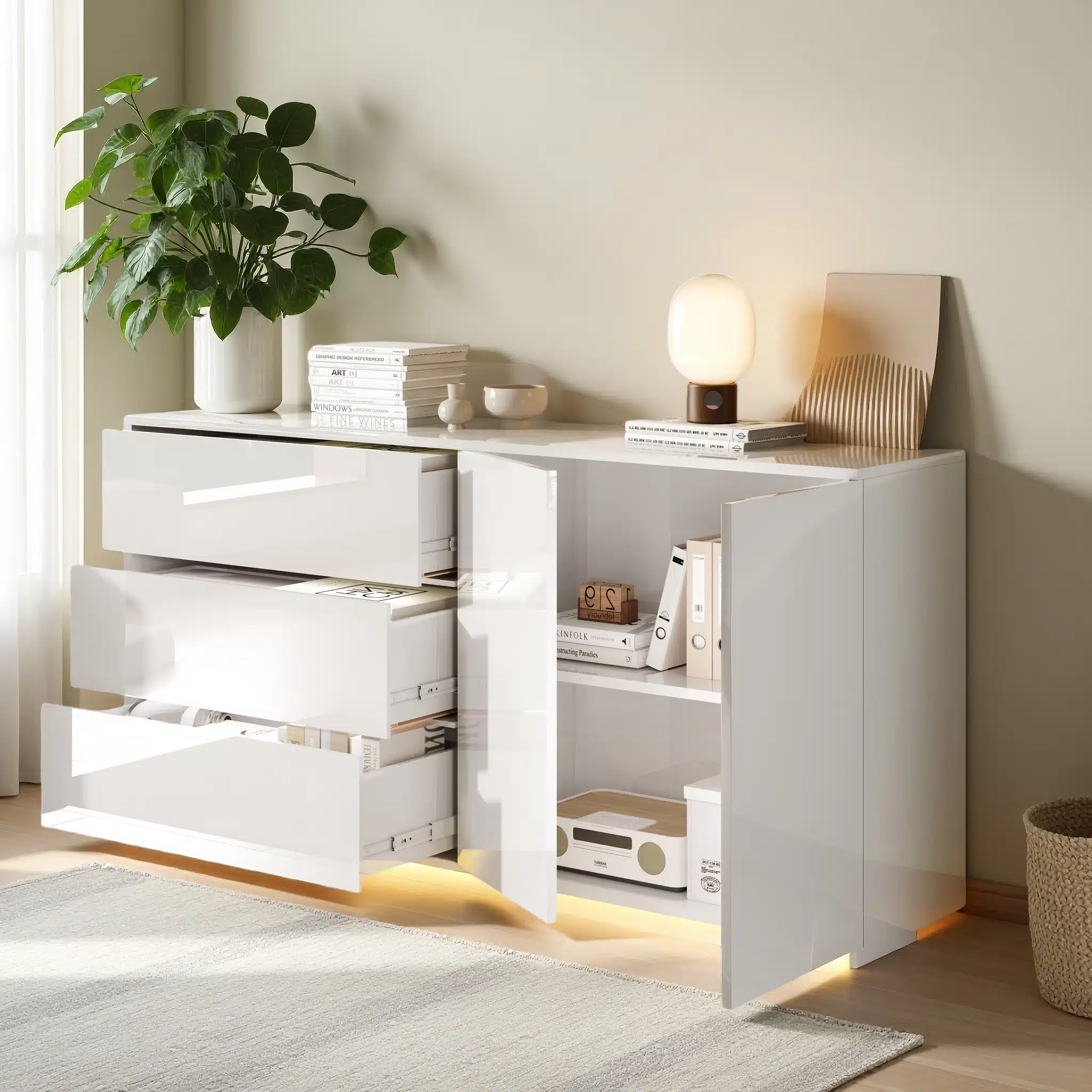 Valge, kaasaegse disainiga kapp LED-valgustusega. Kapp sisaldab kahte uksega sektsiooni ja kolme sahtlit. Puidust põrand ja hele sein panevad eseme esile elutoas. Peal asuvad dekoratiivsed esemed ja taimed lisavad stiili.
