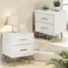 Kahe elegantse valge öökapi komplekt kahe sahtliga ja kuldsete metalljalgadega, paigutatud modernses magamistoas heleda tausta ees. Ühel kapil lamplamp, teisel dekoratiivsed esemed.