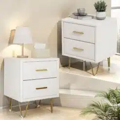 Kahe elegantse valge öökapi komplekt kahe sahtliga ja kuldsete metalljalgadega, paigutatud modernses magamistoas heleda tausta ees. Ühel kapil lamplamp, teisel dekoratiivsed esemed.