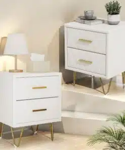 Kahe elegantse valge öökapi komplekt kahe sahtliga ja kuldsete metalljalgadega, paigutatud modernses magamistoas heleda tausta ees. Ühel kapil lamplamp, teisel dekoratiivsed esemed.