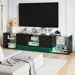 Elegantne TV-alus LED-valgustusega, kaasaegne must disain, kaks keskel asetsevat sahtlit ning läbipaistvad klaasist riiulid, mis pakuvad ruumi hoidmiseks. Sobib ideaalselt stiilsesse elutuppa, pakkudes funktsionaalsust ja kaasaegset esteetikat.