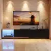 Must TV-alus LED valgustusega, kolm sahtlit, 240x38x44 cm, kaasaegse ja elegantse välimusega, asetseb elutoas heledal puidust põrandal. Sellel on modernne sisustus, sealhulgas päikeseloojangu pilt seinal, minimalistlikud dekoratsioonid ja taimed.