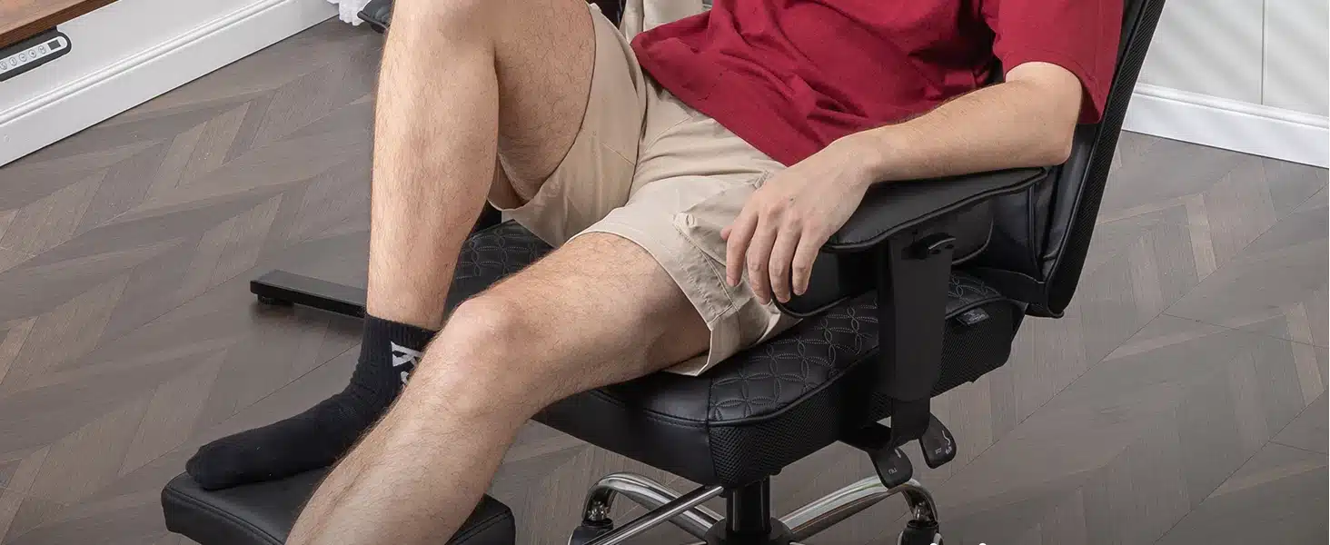 Mees istub mustal vastupidaval ergonoomilisel arvutitoolil, millel on reguleeritav jalatugi, heledal puitpõrandal asuvas toas.
