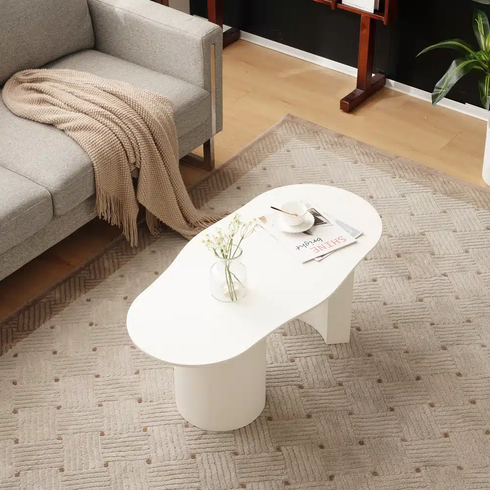 Valge MDF-ga kohvilaud, millel on kaarja disain ja mõõtmetega 100 cm, on paigutatud elegantsele vaibale elutoas, kus on hall diivan ja trendikas beige pleed. Laua peal on puhas valge keraamiline kruus ja väike klaasvaas lilledega.