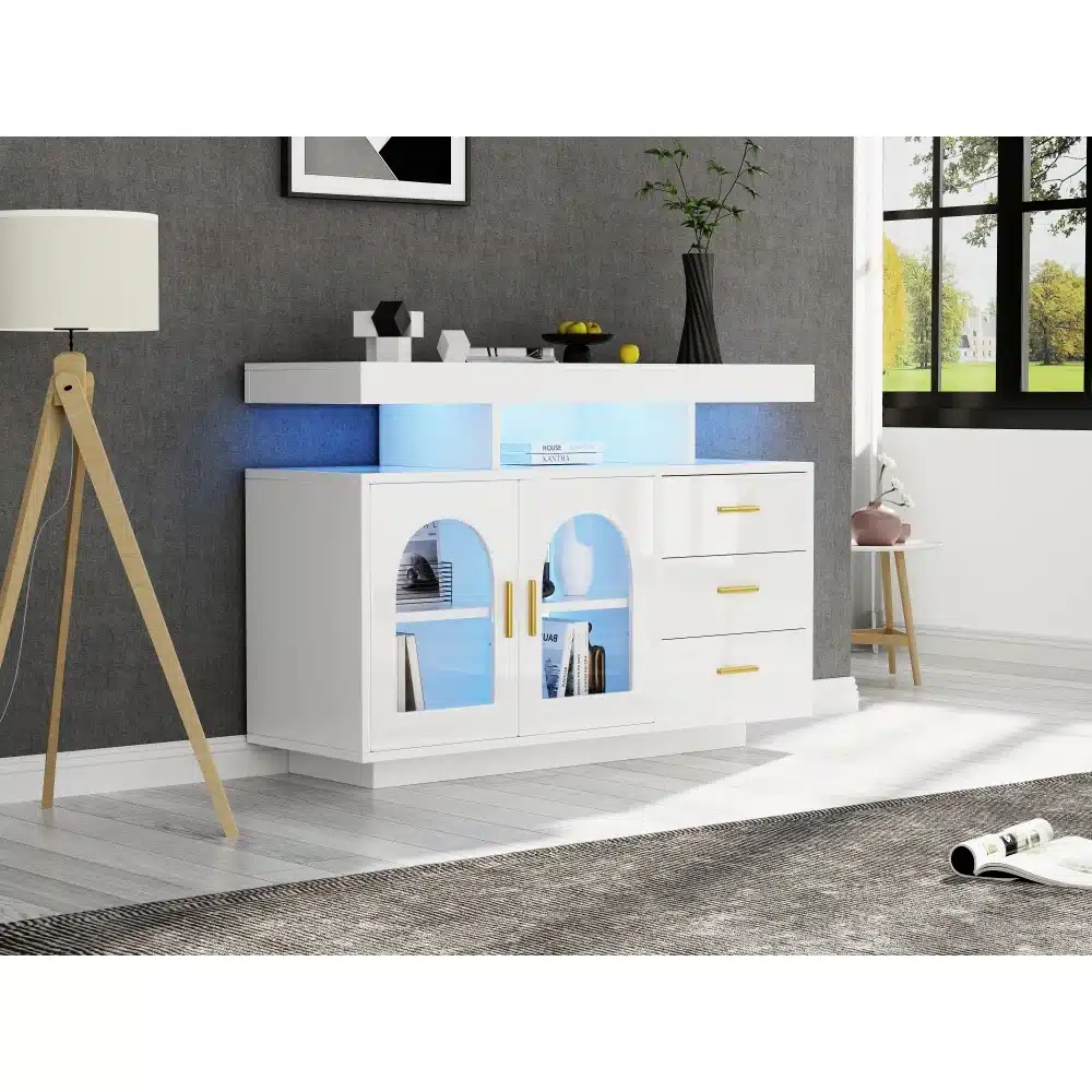 Valge köögikapp LED-valgustusega, kõrgläike viimistlusega ja kolme sahtliga, mis paikneb elegantses kaasaegses elutoas hallide seinte taustal. Kapi peal on dekoratiivsed elemendid ja aknast paistab roheline loodusvaade.