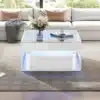 Kaasaegne valge diivanilaud LED-valgustusega ja kahe sahtliga, mille klaaspind peegeldab elegantselt elutoa sisustust, luues hubase atmosfääri.