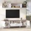 Tv-alus komplekt seinale, 2-osaline, sobib kuni 80-tollisele | E-pood Tootemaailm Elegantselt paigaldatud Tv-alus komplekt seinale, 2-osaline, sobiv kuni 80-tollisele telerile. Komplekt on valget värvi puidu ja avatud riiulitega, mis sobitub hästi helepruuni interjööriga elutoas. Täidetud korvide ja dekoratiivesemetega, paistab päikseline aken taustal.