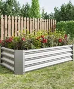 Tõstetud aiapeenar tsingitud terasest mõõtmetega 120x60x30 cm, täis rohelisi taimi ja lilli, paigutatud lopsaka muruga aeda puidust tara ees.