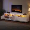 TV-alus LED-valgustusega, valge kõrgläige, 175 cm | E-pood Tootemaailm Valge kõrgläikega TV-alus LED-valgustusega, 175 cm, mis on paigutatud elutuppa. Alusel on avatud uksed, mis paljastavad oranži valgusega valgustatud riiulid, täites ruumi kaasaegse ja sooja atmosfääriga. Taustal on öine taevas suurel ekraanil ning kõrval seisab stiilne lamp koos toataimega.