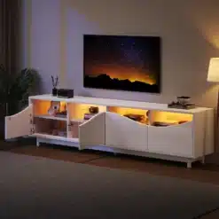 Valge kõrgläikega TV-alus LED-valgustusega, 175 cm, mis on paigutatud elutuppa. Alusel on avatud uksed, mis paljastavad oranži valgusega valgustatud riiulid, täites ruumi kaasaegse ja sooja atmosfääriga. Taustal on öine taevas suurel ekraanil ning kõrval seisab stiilne lamp koos toataimega.