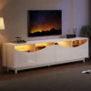 TV-alus LED-valgustusega, valge kõrgläige, 175 cm | E-pood Tootemaailm Valge kõrgläikega TV-alus LED-valgustusega, mõõtmetega 175 cm, modernses elutoas tumedal puidust põrandal. Alus sisaldab mitut riiulit, millel on kollane valgustus, ning sellel on asetatud dekoratiivsed esemed, taustal on lamp ja televiisor.