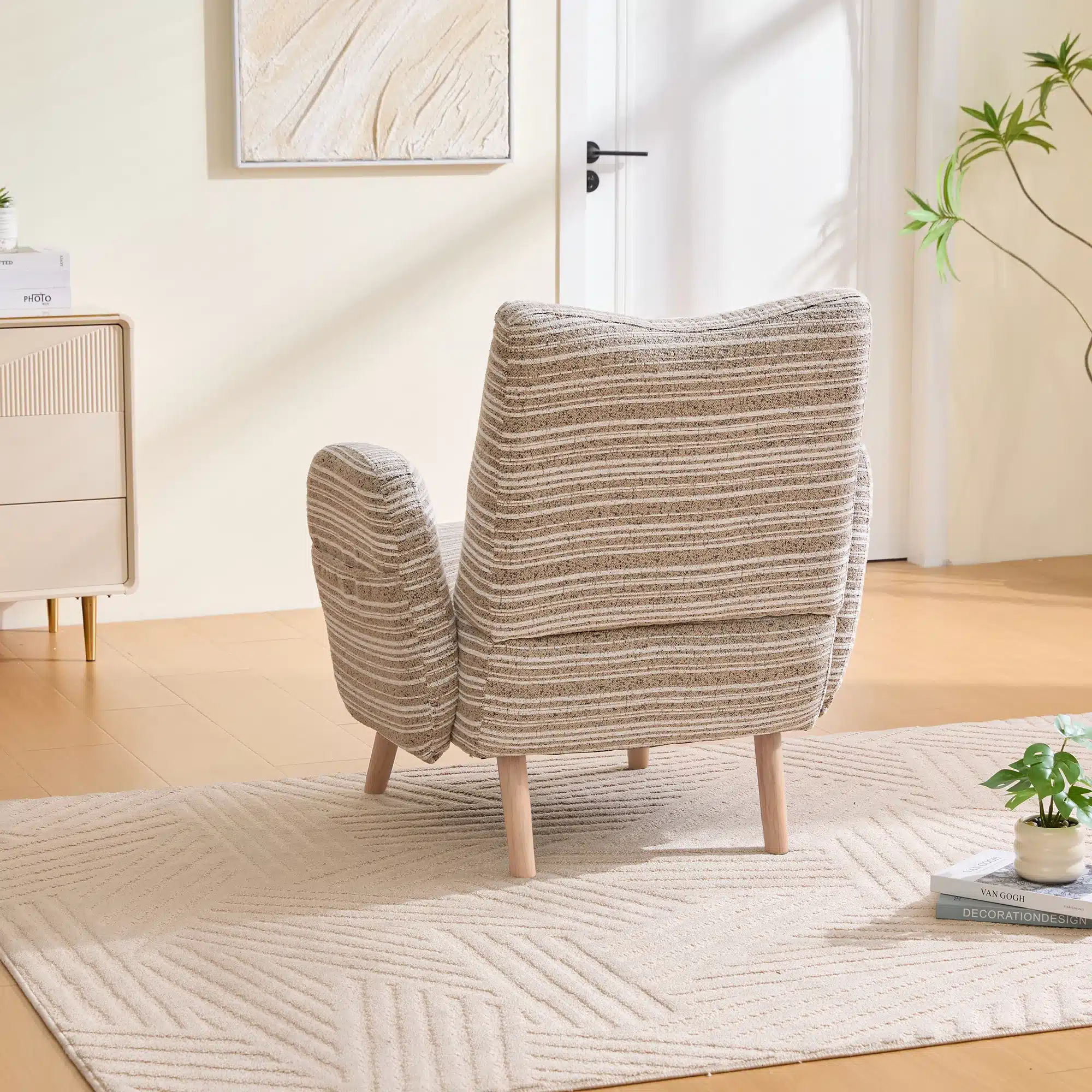 Mugav ja stiilne tugitool küljetaskuga, millel on beež ja hall triibuline kangas ning naturaalsed puidust jalad, seisab heledal vaibal, minimalistlikus elutoas taustal valgus ja roheline taim.