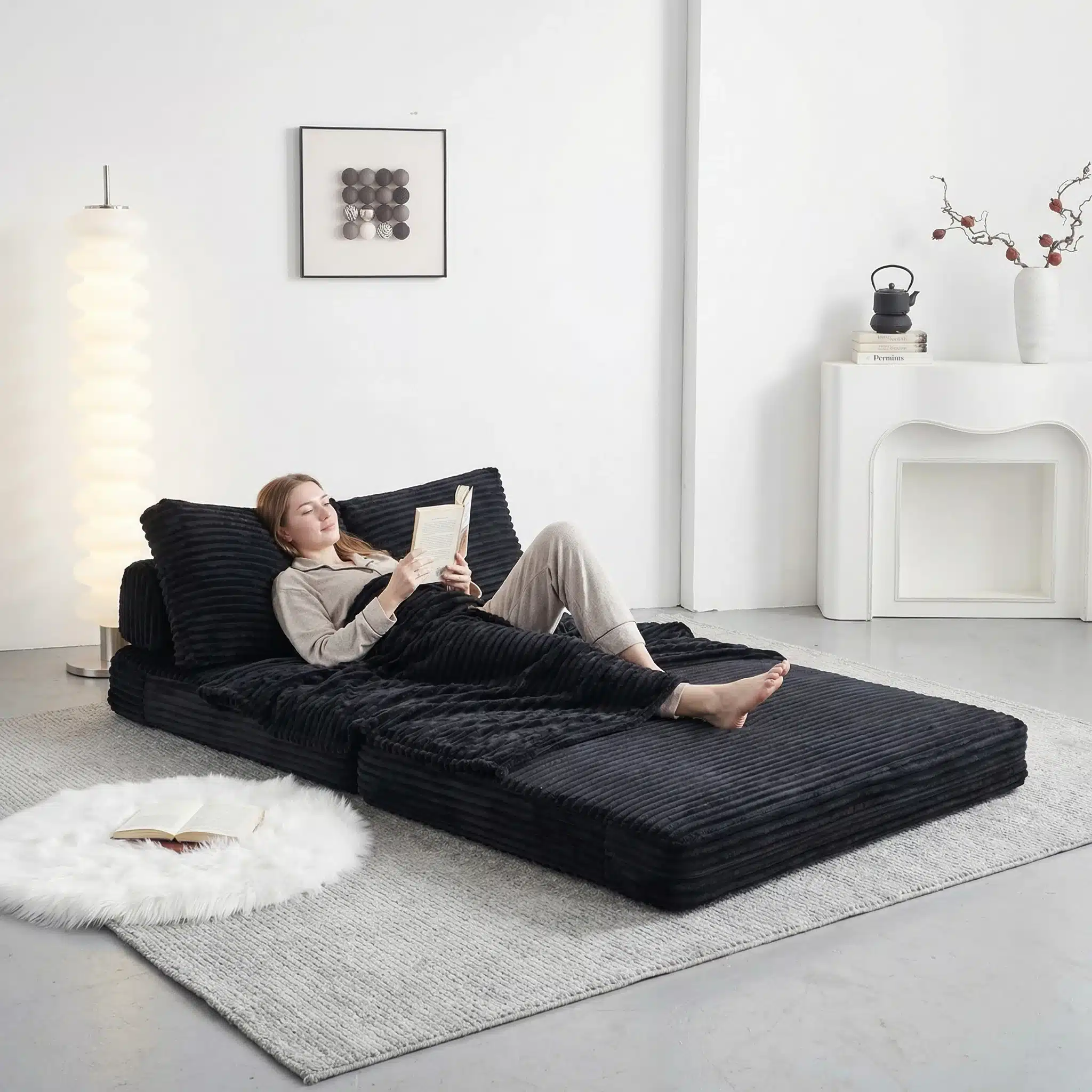 Naine lõõgastub mustal kokkupandaval diivanvoodil, mis on sobilik väikestele ruumidele, asub neutraalses elutoa interjööris heledate seinte ja minimalistliku kaunistusega.