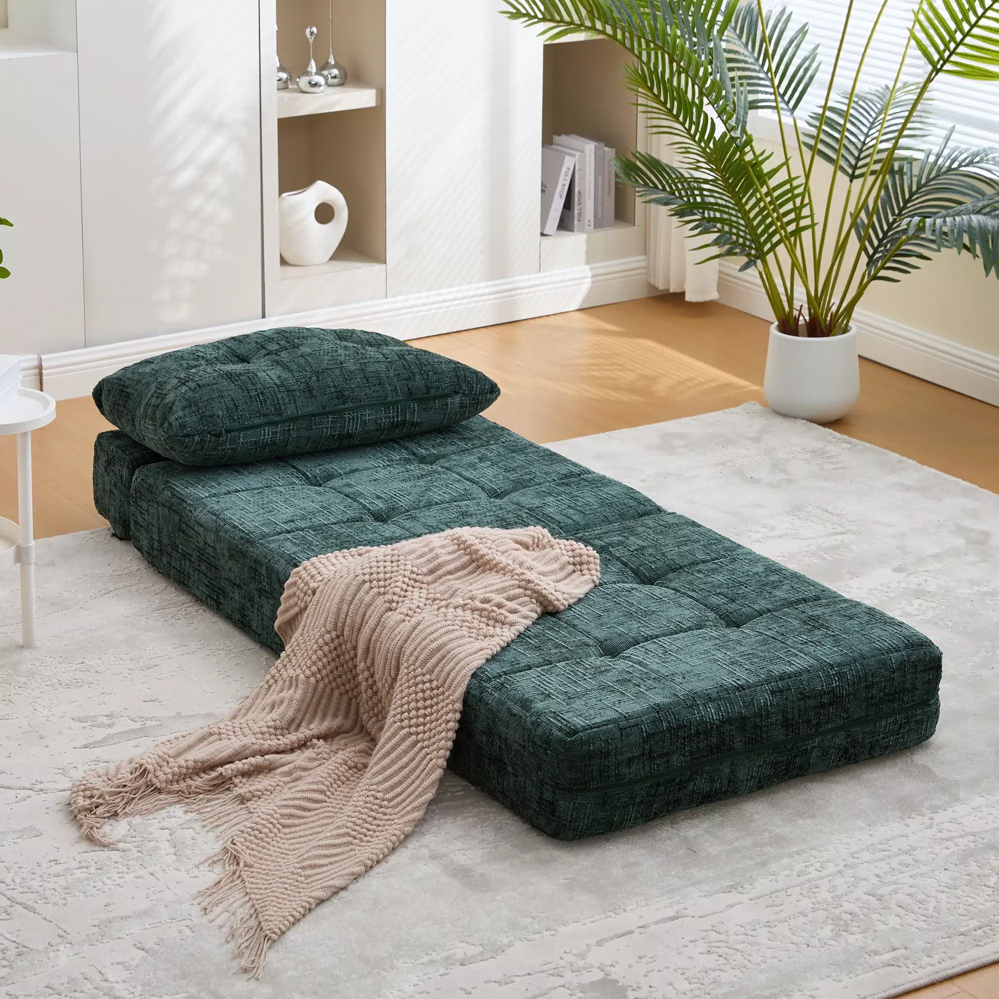 Tumedaroheline volditav ühekohaline diivanvoodi chenille padjaga, millel beige pleed, paikneb helehallil vaibal valges toas, taustal toataim ja riiul.