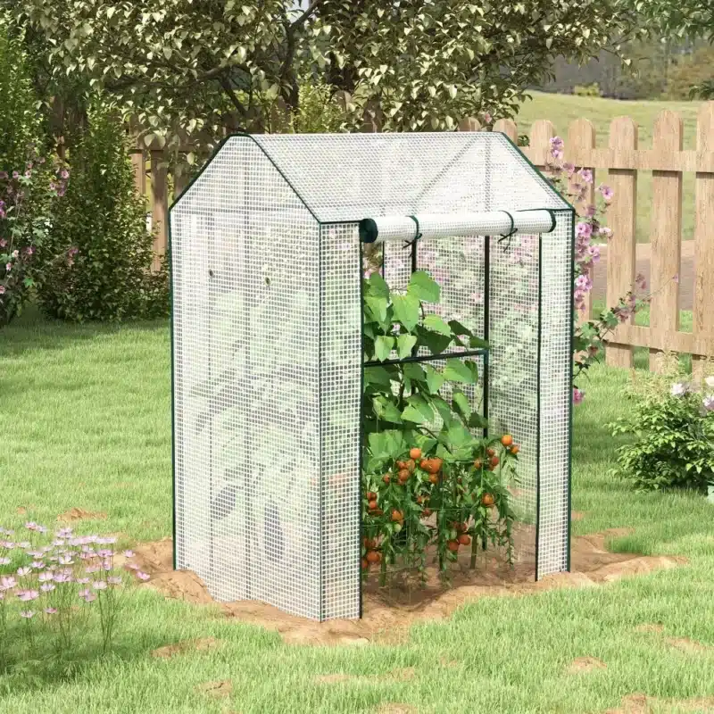 Valge kasvuhoone kahekordse uksega aias, mis on tuulekindel ja mõõtmetega 100x80x150 cm. Kasvuhoone sees kasvavad tomatitaimed, ümbritsetud rohelisest murust ja lilledega.