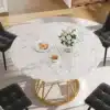Elegantselt kujundatud ümmargune söögilaud, millel on marmorimustriga pind ja kuldsed metalljalad. Laual on asetatud valge vaas valgete lilledega. Tumehallid toolid ümbritsevad laua lihtsas, kuid stiilses interjööris.