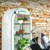 Kasvuhoone ukse ja riiulitega, talvekindel, valge | E-pood Tootemaailm Valge talvekindel kasvuhoone avatud ukse ja mitme riiuliga, täis lilli ja taimi. Taustal telliskivisein, kujutamas hoolitsevat aiapidajat.