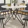 Ümmargune söögilaud marmorviimistlusega, 100x100 cm, koos mustade metalljalgadega, ümbritsetud hallide polsterdatud toolidega, heledal vaibal avaras ja valgusküllases ruumis.