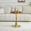 Ümmargune diivanilaud 60 cm, valmistatud pähklipuu MDF-st ja seisab elegantsel kuldsest metallist jalal, valges elutoas beežil vaibal. Laua peal on kaks kuldset dekoratiivset eset.