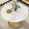 Ümmargune söögilaud marmorist plaadiga ja kuldsete jalgadega, millel on peal vaas ja puuviljad. Laud asetseb heledal vaibal, minimalistlikul taustal.