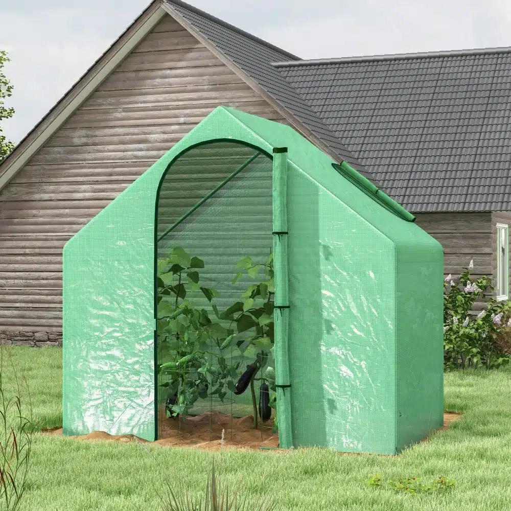 Roheline kasvuhoone aknaga, PVC katte ja vastupidava terasraamiga, mõõtmetega 180x100x168 cm, asub murul esiplaanil vana puumaja taustal.