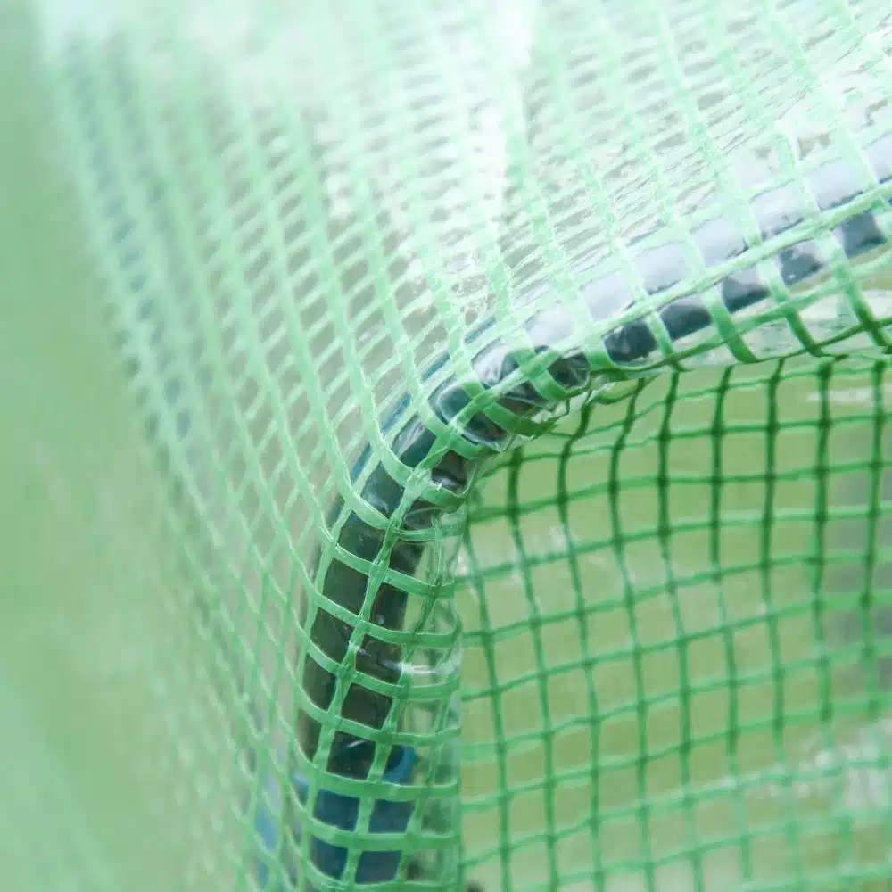 Detailvaade kasvuhoonest aknaga, mis on kaetud tugeva PVC kattega ja toetub vastupidavale terasraamile. Roheline võrgustik lisab kasvuhoonele lisakaitset ja tugevust.