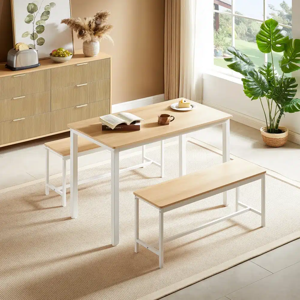 Minimalistlik söögilaud koos pingidega helepruunist puidust, valgete metalljalgadega, asetatud heledale vaibale valgusküllases elutoas. Köögimööbel, taim ja aknast paistev vaade täiustavad kaasaegset interjööri.