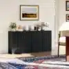 Kapp musta puidu imitatsiooniga, volditud uks | E-pood Tootemaailm Elutoas asuv kapp musta puidu imitatsiooniga, millel on volditud uksed. Kapp on elegantne ja sobib erinevatele tarvikutele, kuna esiplaanil on nõud ja kohvimasin. Taustal valged seinad maalidega.