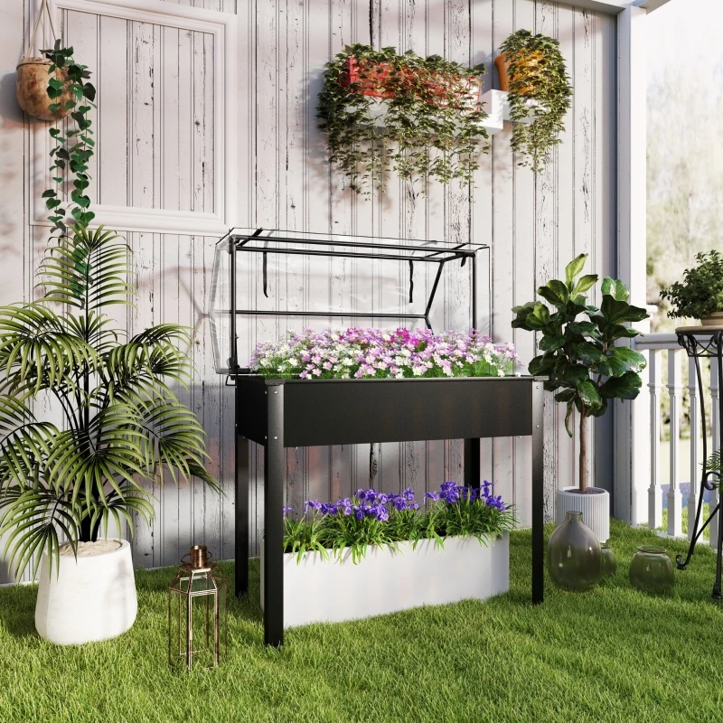 Must tõstetud istutuskast kasvuhoonekaanega mõõtmetega 100x48x124 cm, täidetud roosade ja violetsete lilledega, asetseb rohelisel murul heleda veranda taustal, ümbritsetud erinevate rohetaimedega pottides.