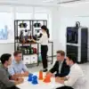 3D printeri alus ratastega kahele printerile metallist | E-pood Tootemaailm Kontoriruum, kus taustal on 3D printeri alus ratastega kahele printerile metallist, millel asetsevad aktiivsed printerid; esiplaanil lauaga, mille taga istuvad neli inimest arutamas 3D prinditud objekte.