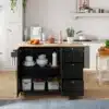 Köögikapp ratastel, kokkupandav, 3 sahtliga, 135 cm | E-pood Tootemaailm Elegantne must köögikapp ratastel, kokkupandav ja 3 sahtliga, 135 cm, paistab helges köögikeskkonnas. Kapil on hele puidust tööpind, millest vasakul on näha veiniklaas ja erinevad tarvikud kappide kõrval. Mitmed rohelised toataimed ja köögitarvikud taustal lisavad mugavust.