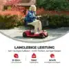 Naine istub punasel elektrilisel neljarattalisel liikumistoolil eakatele, millel on eemaldatav aku, päikesepaistelisel rajal punaste põõsaste ja puumajaga taustal.