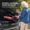 Elektriline neljarattaline liikumistool eakatele, eemaldatav aku | E-pood Tootemaailm Naine paneb kokkuvolditud punast elektrilist neljarattalist liikumistooli auto pagasiruumi, taustal on roheline lehestik ja tellissein.