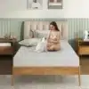 Ortopeediline vahtmadrats 120x200cm 3D õhulise kattega | E-pood Tootemaailm Naine istub hubases magamistoas Ortopeedilisel vahtmadratsil 120x200cm 3D õhulise kattega, hellitades valget kassi. Madratsi neutraalne värv sulandub stiilse puidust voodiraamiga, taustal pehmed padjad ja öökapp.