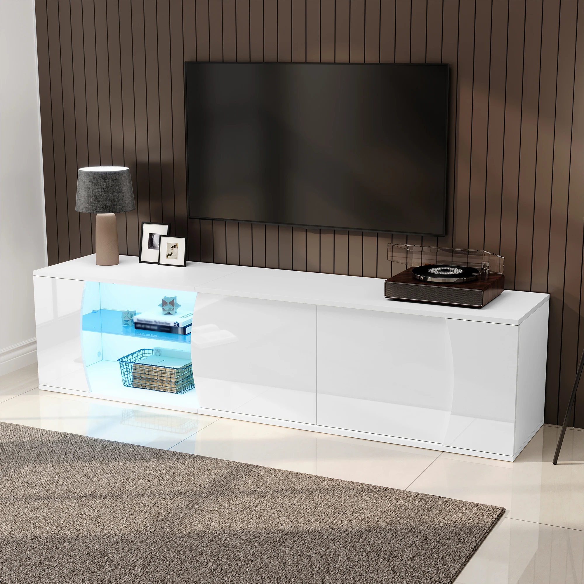 Elegantne TV-alus LED-valgustusega, valmistatud karastatud klaasist, pikkusega 180 cm. Pildil on näha kaasaegne valge alus, millel on sisseehitatud valgustus, minimalistlik disain ning mugavad sektsioonid esemete paigutamiseks. Taustal pruunid seinapaneelid, läheduses pehmel hallil vaibal asetsevad dekoratiivsed esemed.