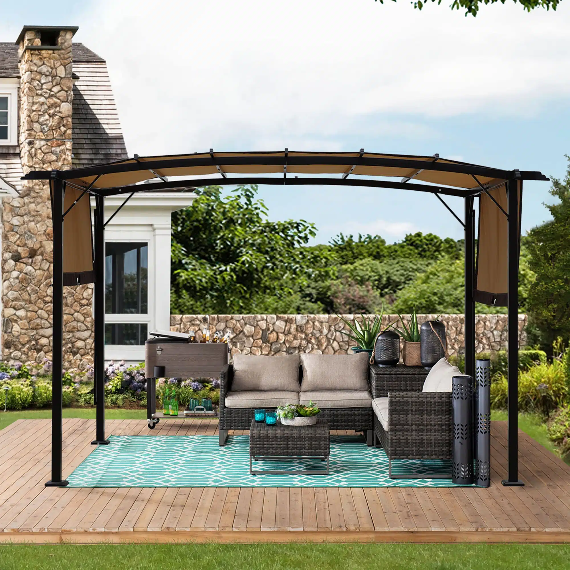 Elegantne Pergola päikesevarir veekindel UV-kaitsega 12x9, mis on paigaldatud puidust terrassile. Pergola kattestruktuur on must metallist, mille kohal on beež veekindel kate. Taustal on lopsakas roheline aed ja kivimaja.