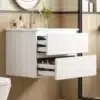 Valge vannitoakapp LED valgusti ja integreeritud valamuga, mille sahtlid on avatud. Taustal hele sein ja mõned esemed valamu peal.