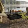 Kokkupandav matkalaud 120x55cm kerge hoiukotiga | E-pood Tootemaailm Kokkupandav matkalaud 120x55cm kerge hoiukotiga telkimisplatsil, laual on kohvikann, kruus, leib, maasikad ja puuviljad, taustal telk ja järv.