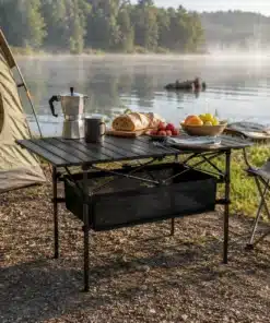 Kokkupandav matkalaud 120x55cm kerge hoiukotiga telkimisplatsil, laual on kohvikann, kruus, leib, maasikad ja puuviljad, taustal telk ja järv.