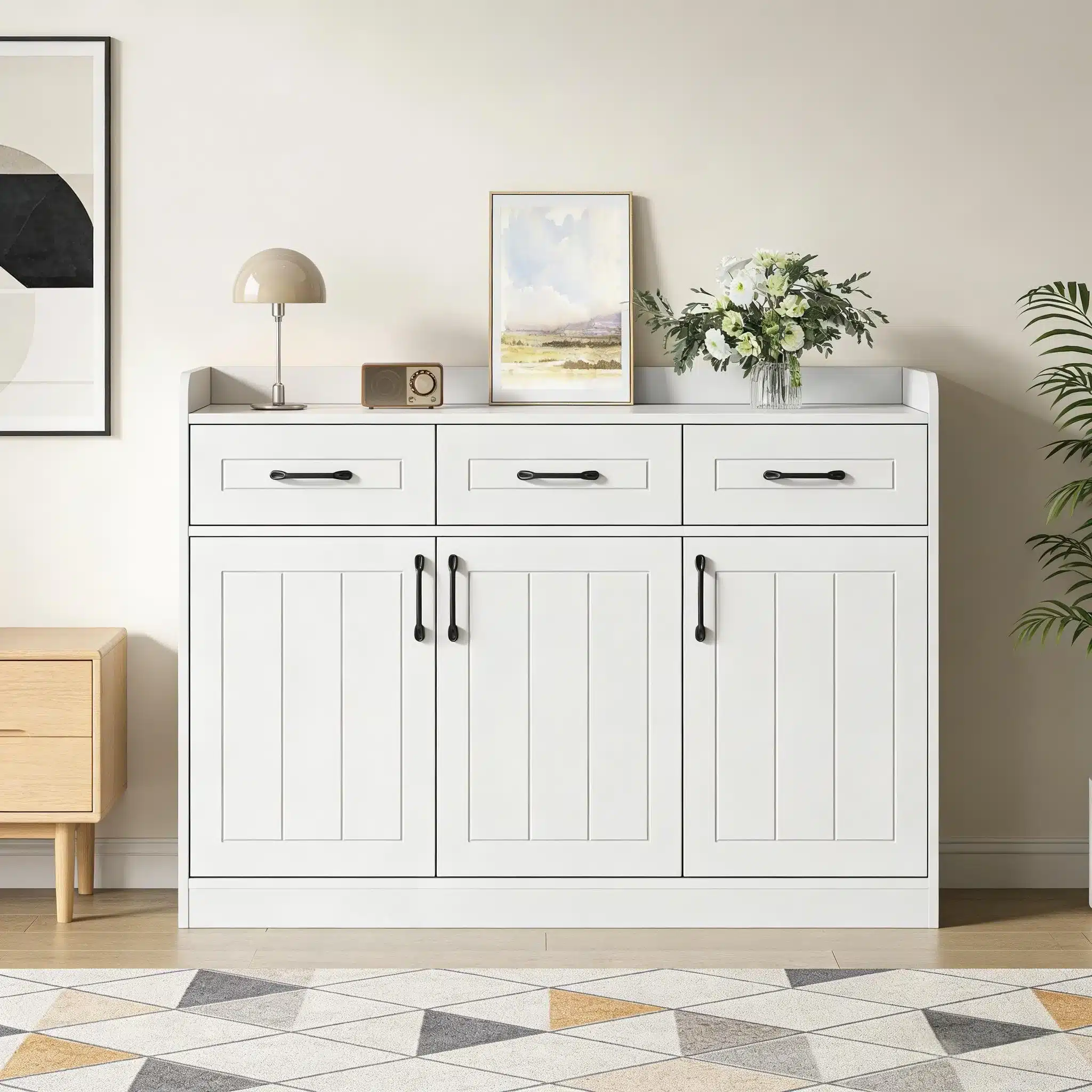 Valge kummut triibulise disainiga, mõõtmetega 120x35x80 cm, millel on kolm sahtlit, asub ruumis heleda seina ja kunstipõrandal koos dekoratiivsete elementidega, nagu lamp ja lillevaas.