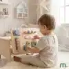 Montessori tegevustahvel lastele peenmotoorika arendamiseks | E-pood Tootemaailm Väikelaps mängib puidust tegevuslaua ehk busy boardiga heledas lastetoas