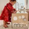 Montessori tegeluskuubik lastele 12+ kuud | E-pood Tootemaailm Beebitüdruk punases ruudulises kleidis seisab puidust tegevuskeskuse kõrval